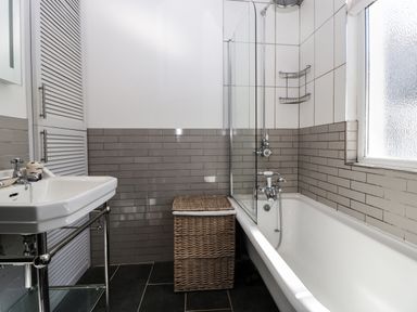 A bathroom with a white bathtub a glass shower screen a wicker laundry basket a white sink and tiled walls at Ferriwais in Appledore