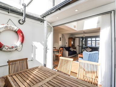 A wooden table and chairs on a patio with a life ring on the wall and a living room with sofas and a cupboard at Ferriwais Appledore