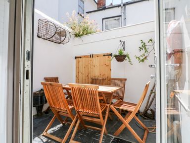 An outdoor patio with a wooden table and six wooden chairs against white walls with hanging plants at Ferriwais in Appledore