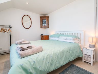 A bedroom with a double bed covered by a green patterned duvet with towels on it a wooden corner cabinet a white bedside table with a lamp and a rack with wooden hangers at Ferriwais in Appledore