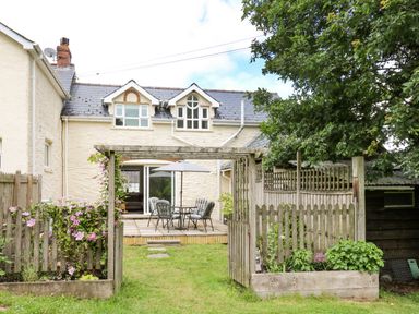 A garden with a wooden pergola and flower beds leading to a patio with a table, chairs, and an umbrella at Spring Cottage in Pentre-Cwrt