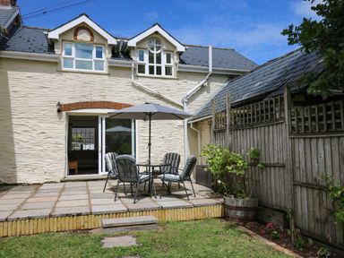 An outdoor patio with a table umbrella and chairs next to a white stone house with windows and a wooden fence at Spring Cottage in Pentre-Cwrt