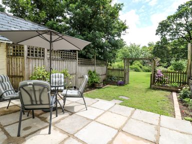 An outdoor patio with cushioned chairs around a table under an umbrella and a garden with a wooden fence and gate at Spring Cottage in Pentre-Cwrt