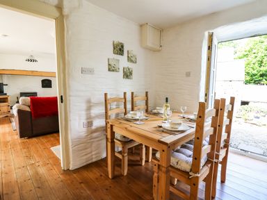 A dining area with wooden table and chairs set for four next to an open door and a living room visible through a doorway at Spring Cottage in Pentre-Cwrt