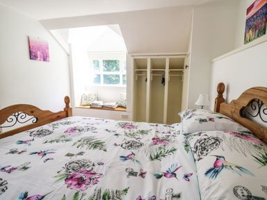 A bedroom with a floral patterned bedspread wooden bed frame open wardrobe window seat with cushions and a lamp at Spring Cottage in Pentre-Cwrt