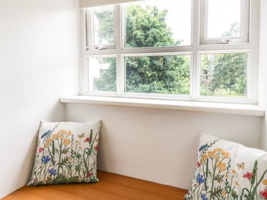 A window seat with two floral patterned cushions under a white window with green trees outside at Spring Cottage in Pentre-Cwrt