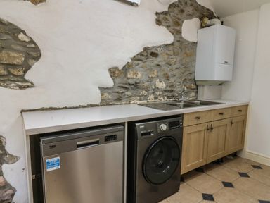 A kitchen area with a dishwasher washing machine wooden cabinets and a double sink under a stone and plaster wall at Spring Cottage in Pentre-Cwrt