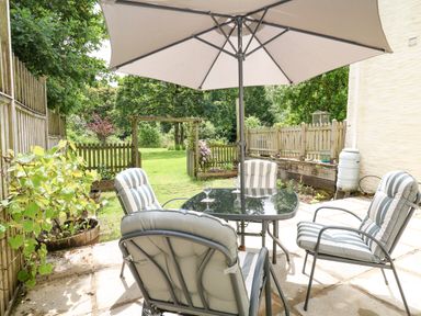 A patio with a glass table and four cushioned chairs under a large umbrella next to a garden with grass and trees at Spring Cottage in Pentre-Cwrt