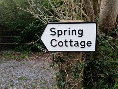 A white directional sign with black text reading Spring Cottage attached to a wooden post surrounded by plants
