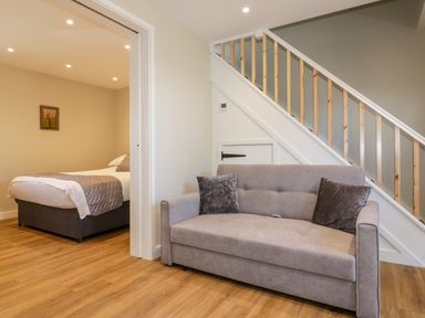 A room with a gray sofa under stairs and a bedroom with a bed visible through an open doorway at The Granary in Helland near Bodmin