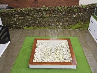 A small water fountain with white stones and a wooden frame on a green artificial grass area in a backyard at Crowthers in Dartmouth