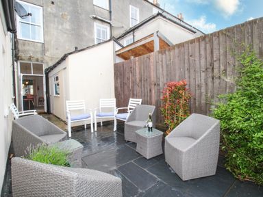 A small outdoor patio area with gray wicker chairs and white chairs next to a wooden fence at Seaside House in Weymouth
