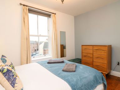 A bedroom with a bed covered in a blue blanket and two folded towels next to a wooden dresser and a window with curtains at Seaside House in Weymouth