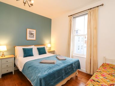A bedroom with a double bed with blue bedding and two towels two bedside tables with lamps a window with curtains and a single bed with floral bedding at Seaside House in Weymouth