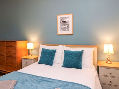 A bedroom with a double bed with teal pillows and a blue throw blanket two bedside tables with lamps and a wooden dresser at Seaside House in Weymouth