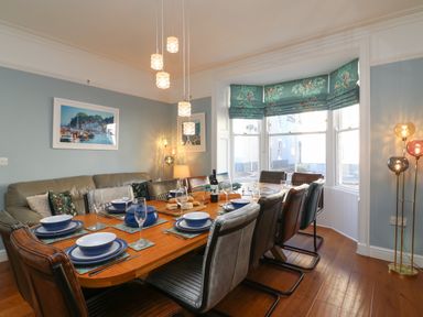 A dining room with a wooden table set for eight people with leather chairs and a bay window with patterned blinds at Seaside House in Weymouth