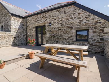 An outdoor patio with a wooden picnic table and potted plants next to a stone building with a glass door and window at The Hayloft in Helland near Bodmin