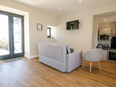 A living area with a gray sofa, a gray armchair, a wall-mounted TV, a clock, and a kitchen with appliances at The Hayloft in Helland near Bodmin