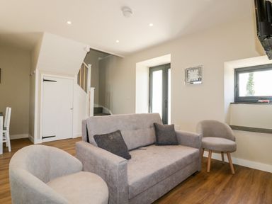 A living room with a gray sofa and two round chairs near a staircase and windows at The Hayloft in Helland near Bodmin