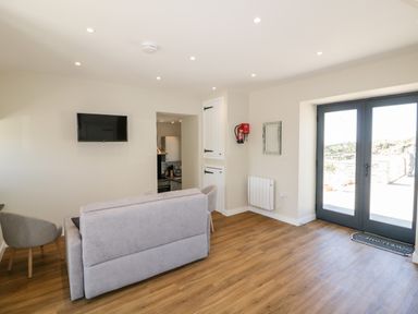 A room with a gray sofa, armchair, wall-mounted TV, double glass doors, wooden floor, and a kitchen visible through an open doorway at The Hayloft in Helland near Bodmin
