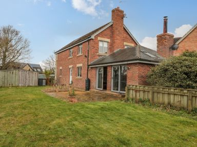 A backyard with a grass lawn a patio and a brick house with large windows and a wooden fence at Ridley Lodge in West Thirston felton
