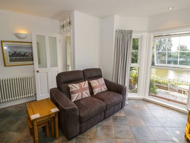 A living room with a brown sofa and two Union Jack pillows next to a wooden nesting table and a bay window overlooking a patio and water at Florence Villas in Worcester