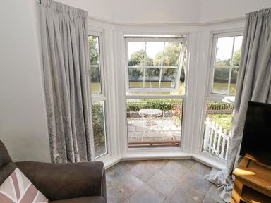 A corner of a living room with a brown armchair and a wooden TV stand near bay windows looking out to a patio with white metal table and chairs at Florence Villas in Worcester