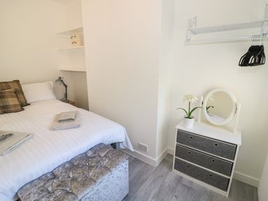 A bedroom with a bed, pillows, folded towels, bedside lamp, shelving, a small dresser with fabric drawers, a mirror, a potted plant and a wall-mounted rack with hangers at Florence Villas in Worcester