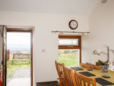 A dining area with a table covered by a yellow checkered tablecloth and wooden chairs next to a window and an open door showing an outdoor bench at Penrallt Fawr in Rhosgadfan near Waunfawr