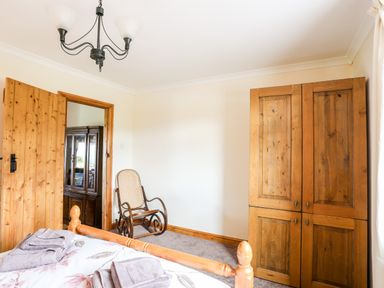 A bedroom with a wooden bed, rocking chair, wooden wardrobe, and an open wooden door at Penrallt Fawr in Rhosgadfan near Waunfawr