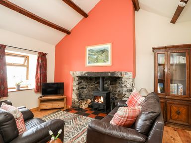 A living room with a stone fireplace and wood burning stove two leather sofas a wooden cabinet a television and a window with red curtains at Penrallt Fawr in Rhosgadfan near Waunfawr