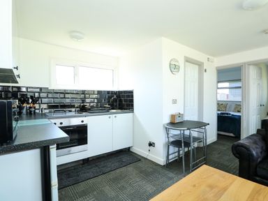 A kitchen with black tile backsplash and white cabinets next to a small dining table with chairs and two doorways at 57 Cherry Park in Chapel St Leonards