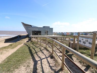 A building labeled North Sea Observatory by the beach with a wooden fence and colorful beach huts in the background at 57 Cherry Park in Chapel St Leonards