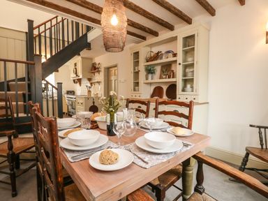 A dining room with a wooden table set for a meal with bread and glassware and a kitchen and staircase in the background at Norden House in Maiden Newton