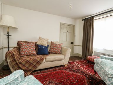 A living room with a beige sofa covered by a plaid blanket and cushions a floor lamp a red patterned rug and a window with curtains at Norden House in Maiden Newton
