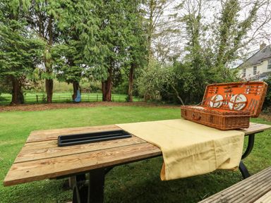 A wooden picnic table with a yellow cloth and a picnic basket set in a garden with trees and a house visible in the background at Norden House in Maiden Newton