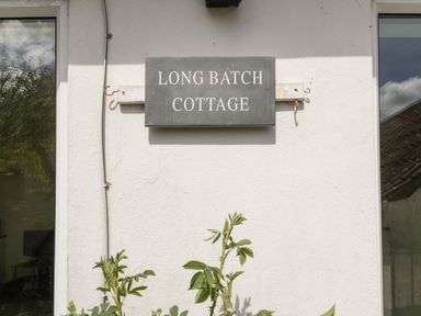 A black sign with white text saying Long Batch Cottage on a white wall between two windows at Long Batch Cottage in Ditcheat