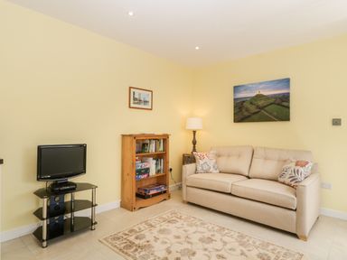 A living room with a beige sofa a wooden bookshelf a floor lamp a small TV on a black stand and two pictures on yellow walls at Long Batch Cottage in Ditcheat