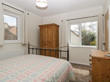 A bedroom with a bed covered by a patterned blanket a wooden wardrobe and a wooden dresser beside windows with curtains at Long Batch Cottage in Ditcheat