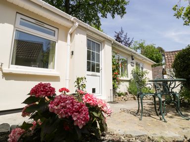 A patio area with pink flowers a white door windows and a green metal table and chairs at Long Batch Cottage in Ditcheat