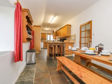 A kitchen with wooden cabinets stools a dining table with cups and plates and red curtains at South Boduel Farmhouse in Liskeard