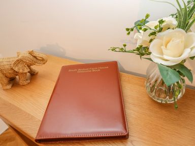 A wooden table with a brown welcome book a wicker elephant figurine and a glass vase with white flowers at South Boduel Farmhouse in Liskeard