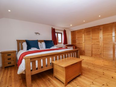 A bedroom with a wooden bed and chest next to a window with red curtains and wooden wardrobes at South Boduel Farmhouse in Liskeard