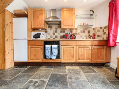 A kitchen with wooden cabinets a microwave a refrigerator an oven with oven mitts a toaster and kettle on the counter and red curtains at Keepers Cottage in Liskeard