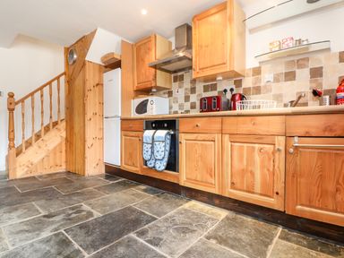 A kitchen with wooden cabinets a tiled floor a white refrigerator a microwave and a staircase at Keepers Cottage in Liskeard