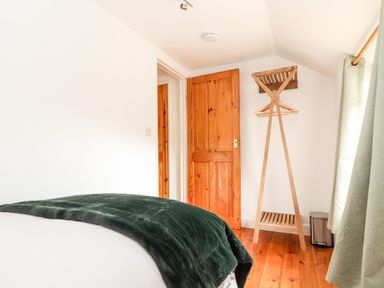 A bedroom corner with a bed covered with a dark blanket an open wooden door a wooden clothes rack with hangers and green curtains at Keepers Cottage in Liskeard