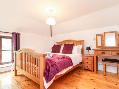 A bedroom with a wooden bed with purple bedding and matching pillows next to a wooden dressing table and bedside cabinet at Keepers Cottage in Liskeard