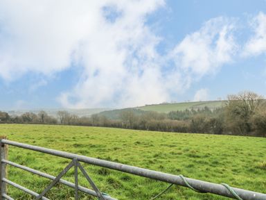 A metal gate opening to a green grassy field with trees and hills under a cloudy sky at Keepers Cottage Liskeard