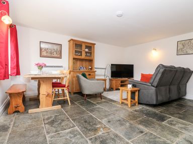 A living room with wooden dining table chairs grey armchairs television and wooden cabinets at Keepers Cottage in Liskeard