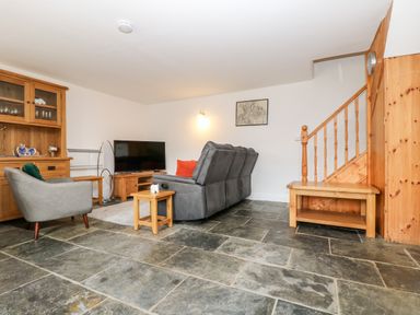 A living room with a gray sofa a television wooden cabinets and stone tile flooring at Keepers Cottage in Liskeard
